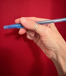 VK Holding Ballpoint Pen A woman's hand holding a blue ballpoint pen with the cap on, against a red background.