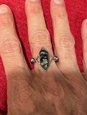 CONQUERing A photo of a woman's hand, showing a ring on the middle finger. The stone is a vertical rectangle, pointed at each end. The stone is black with swirls of foggy grey. The ring itself is stainless steel in a silver color.