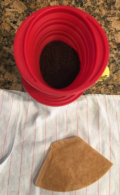 Photo of a red, silicon coffee filter containing coffee grounds. Below it, sitting on a white dish towel with thin red stripes, is a brown, paper coffee filter.