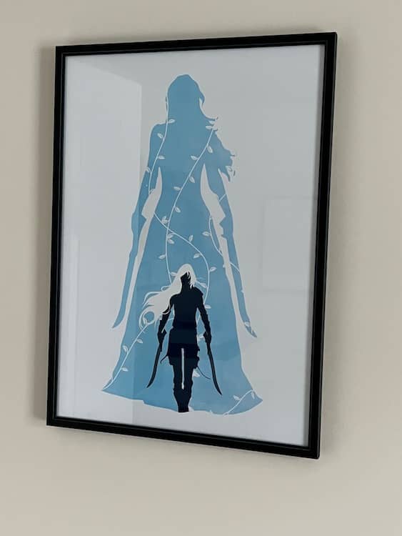 Framed Celaena Artwork showing in light blue a woman with long hair, her back to the viewer, holding two swords at her sides. Inside her dress is an image of the same woman in dark blue, with white hair, stalking toward the viewer wearing fighting leathers and carrying the same swords.