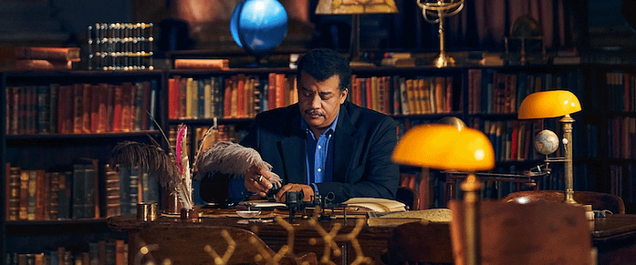 A photo of Neil deGrasse Tyson. He is seated at a desk with bookshelves behind him. He is looking down. He is holding a quill pen and the feathers drape over his hand. He is wearing a dark blue jacket and a medium blue shirt.