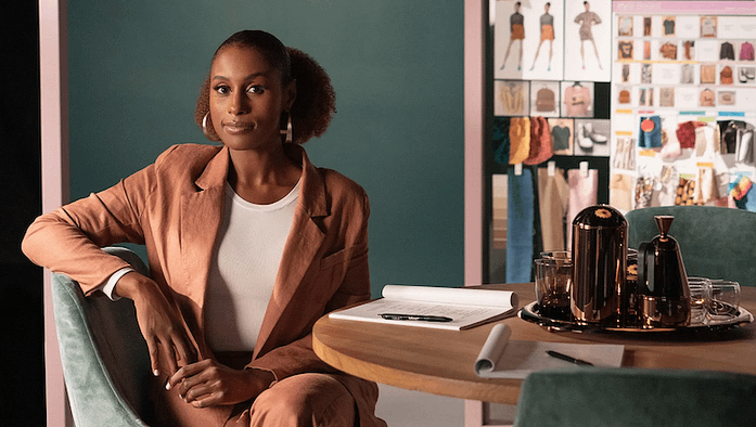 Issa Rae Masterclass A photo of Issa Rae. She is facing the camera while sitting sideways on a chair. Her elbow is bent and on the back of the chair. There is a chalkboard covered in sketches to the back right and a table in the front right with a script and some decorative bottles on top.