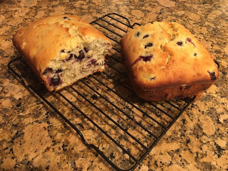 Photo of a loaf of lemon blueberry bread cut in half, sitting on a wire cooling rack.