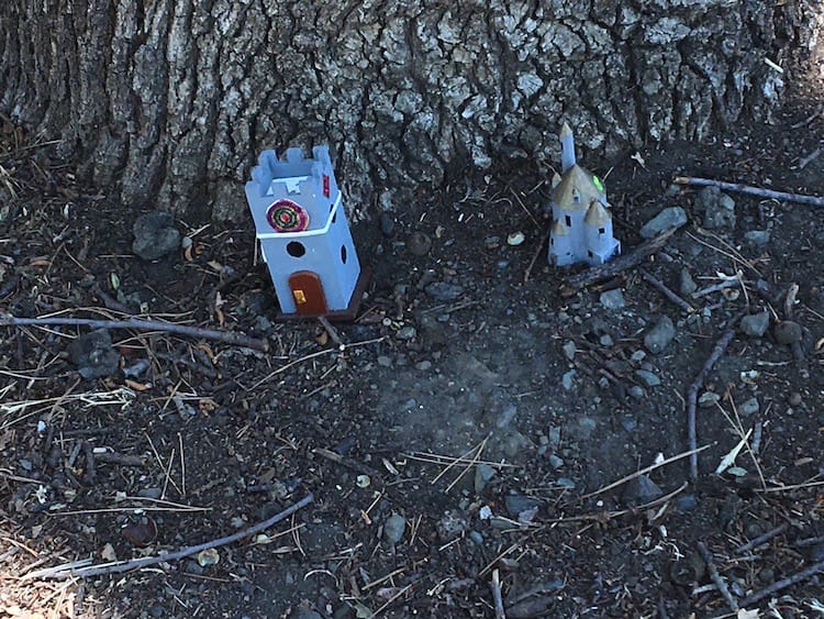 Tower and castle Photo of a small, square gray tower with a circular crest on it and a brown door. About a foot to the right is a gray castle with gold conical roofs on each section. These buildings are set on the ground at the base of a tree.