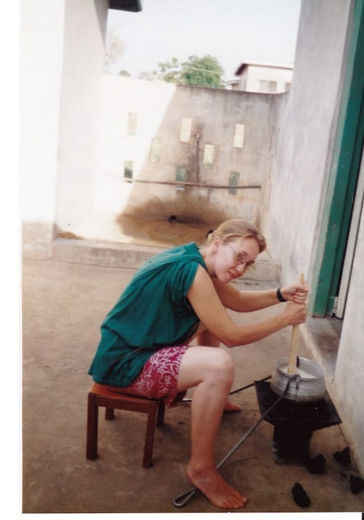 Africa_banku Photo of Vanessa Kier sitting on a stool in front of an unlit brazier on which sits a metal cooking pot. She is stirring the contents of the pot with a wooden spoon.