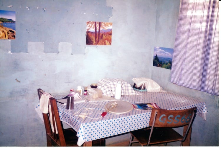 Africa_dining_room2 Photo of a blue and white checkered plastic table cloth on a table with two mismatched wooden chairs. To the right are blue checkered curtains over a window.