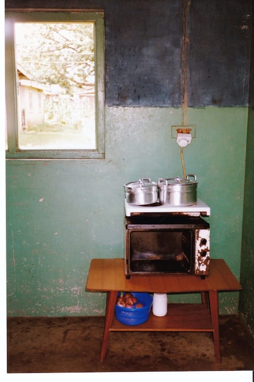 Africa_kitchen2 Photo of a low wooden table with a shelf beneath containing a blue plastic container holding onions. On top of the table is an old, rusted electric oven with two burners on top. Two aluminum cooking pots with handles are on the burners.