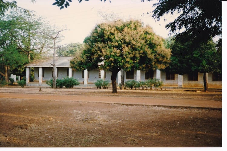 Africa_school1 Photo of dirt in the foreground with a few trees and a row of low bushes in front of a white, single story building with square columns.