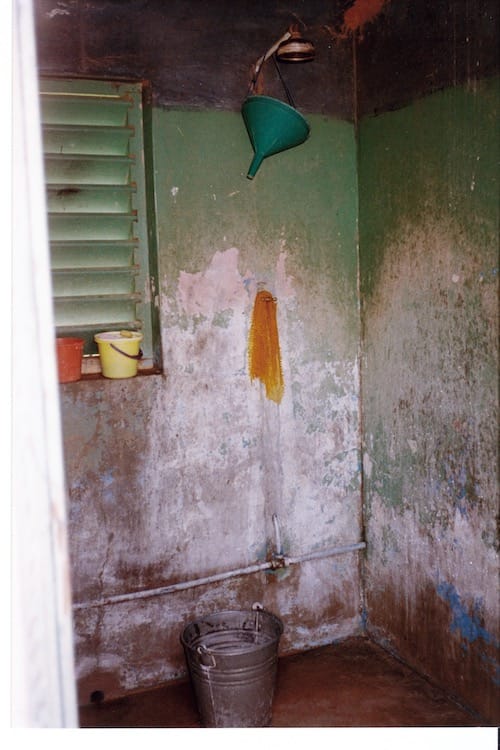 Africa_shower Photo of a corner of a bathing room with a five gallon metal bucket underneath a showerhead. Walls are missing part of their green paint.