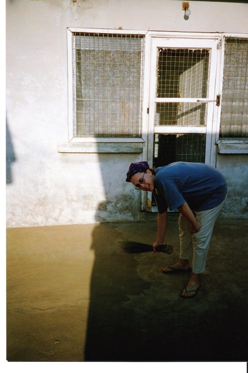 Africa_sweeping Photo of a courtyard with the left half in the sun and the right half in shade. Vanessa Kier is bent over, holding a short rush broom as she sweeps.